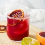 A glass of vibrant Blood Orange Margarita Mocktail with ice and a blood orange slice, next to a halved lime and blood orange.