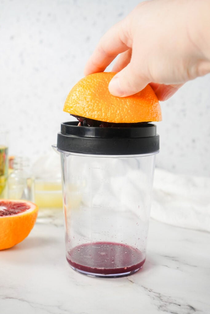 Hand pressing half a blood orange on a juicer over a clear container, with vibrant juice collecting at the bottom&mdash;perfect for your next mocktail or margarita mocktail.