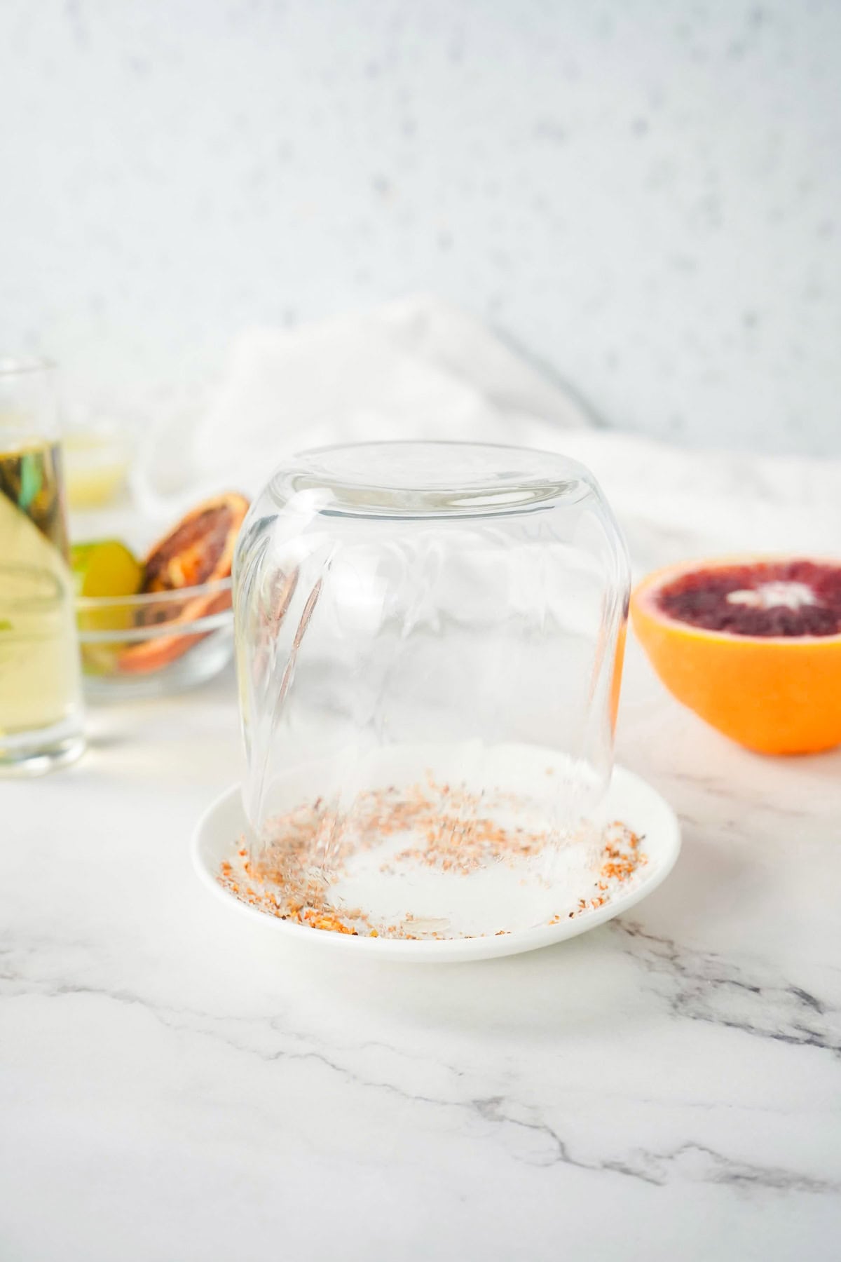 An upside-down glass rim being dipped into a plate of seasoning, with citrus and drinks in the background&mdash;perfect for crafting a refreshing Margarita Mocktail.