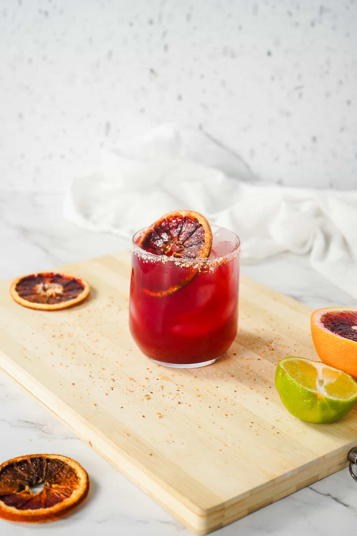 A vibrant red Blood Orange Margarita Mocktail garnished with a blood orange slice, served on a wooden board surrounded by fresh citrus fruits.