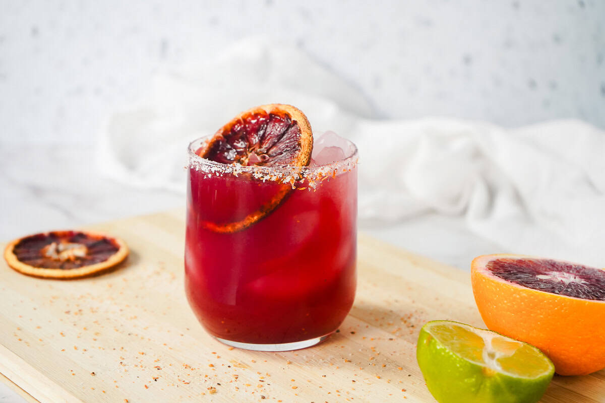 A glass of Blood Orange Margarita Mocktail with ice, garnished with a blood orange slice, sits next to fresh lime and orange halves.