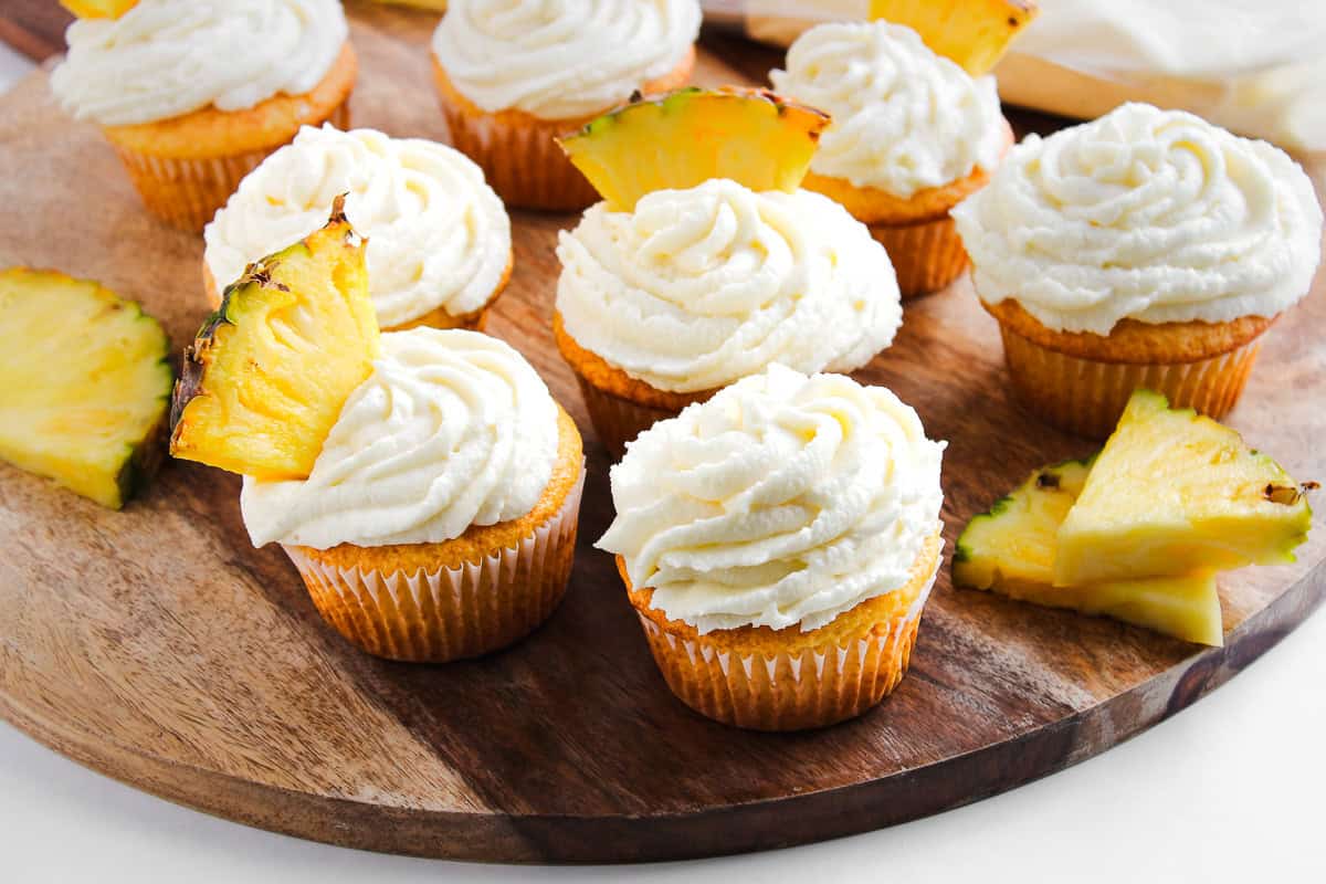 Dole Whip Cupcakes with white frosting and pineapple slices on top, arranged on a wooden board.
