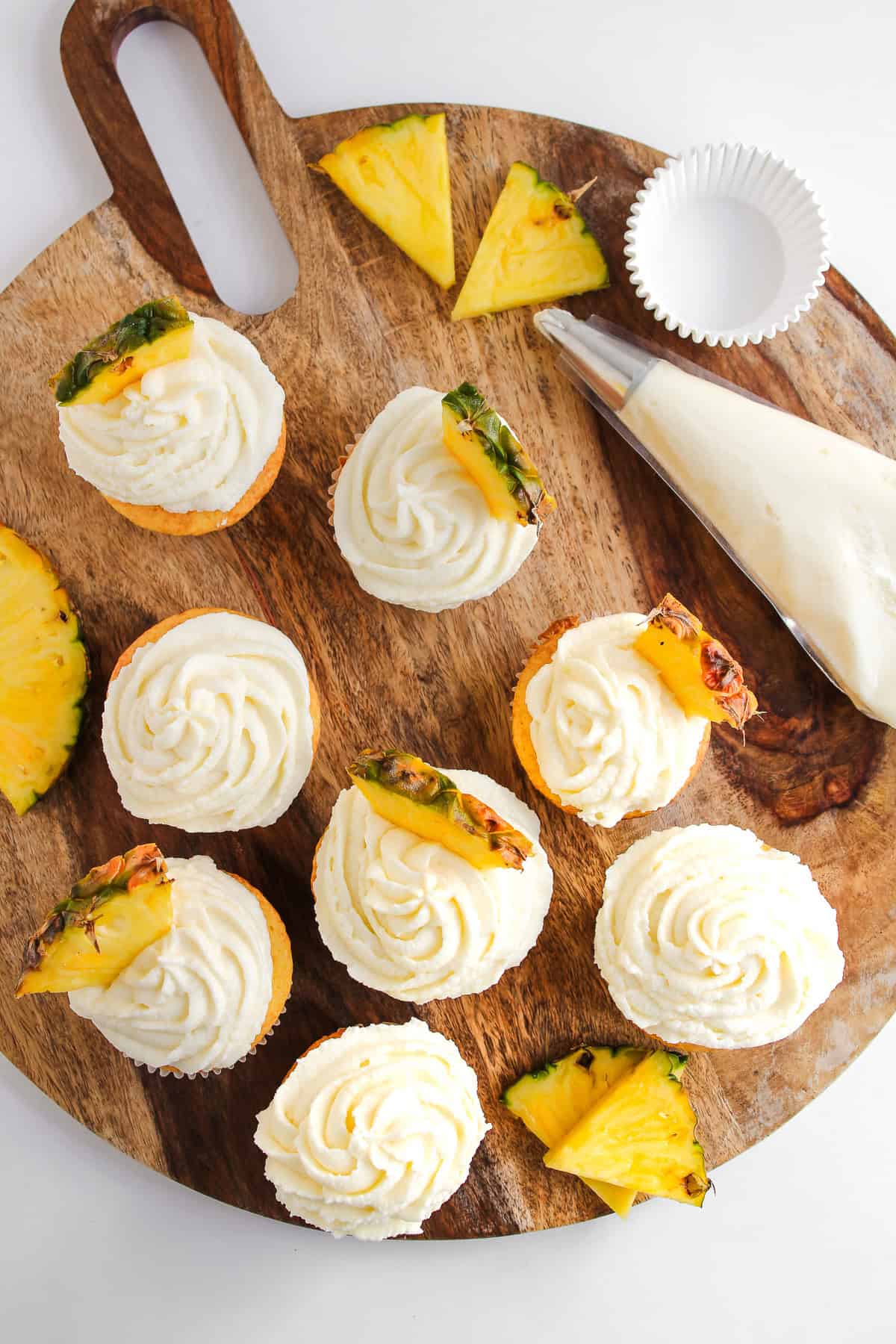 Vanilla Dole Whip Cupcakes with swirled frosting and pineapple slices on a wooden board, accompanied by a piping bag and cupcake liner.