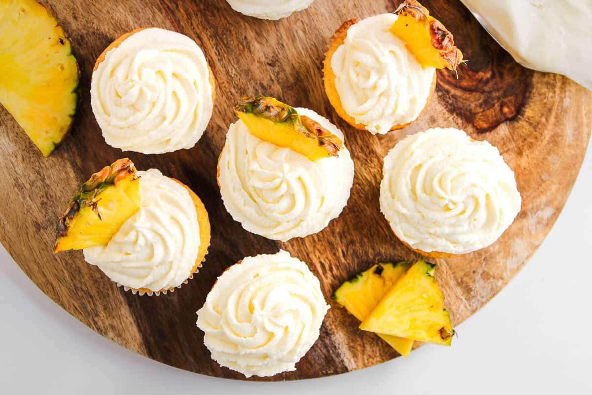 Dole Whip Cupcakes with swirled white frosting and pineapple slices are artfully arranged on a round wooden board.
