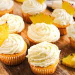 Dole Whip Cupcakes with swirled white frosting, each topped with a pineapple slice, are arranged on a wooden board.