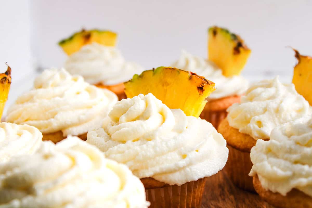 Vanilla Dole Whip Cupcakes with swirled white frosting, each topped with a slice of pineapple, arranged on a wooden surface.