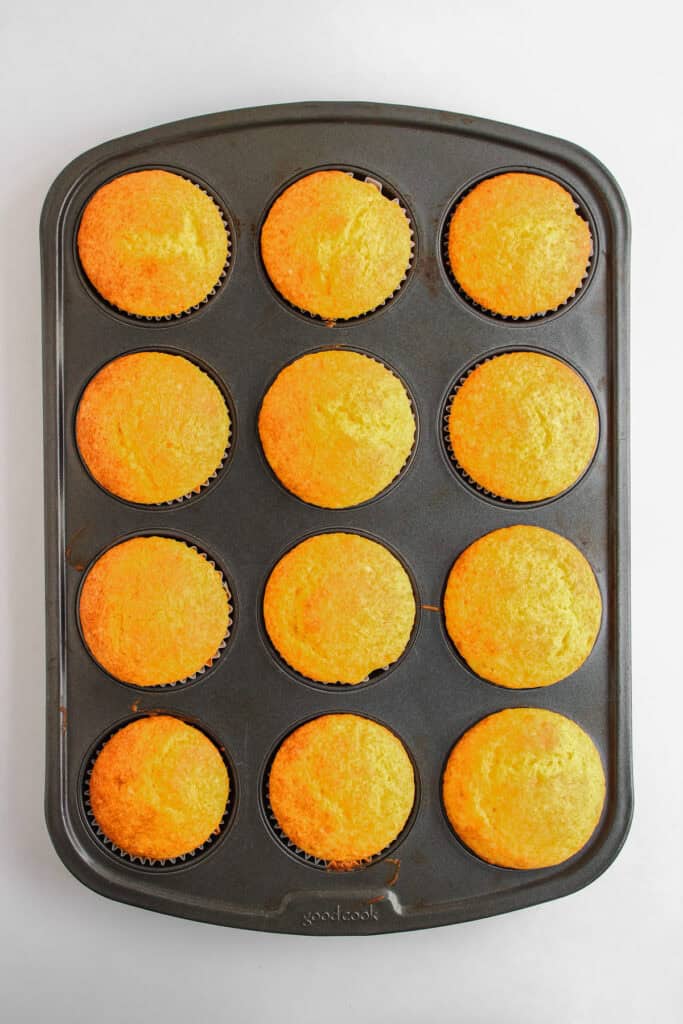 A muffin tin with twelve freshly baked golden Dole Whip Cupcakes in brown paper liners sits on a white background.