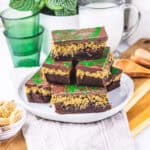 A plate of chocolate layered dessert bars with a green swirl on top, made using a rich Dubai Chocolate Brownie recipe, sits next to a glass of milk and green glasses.