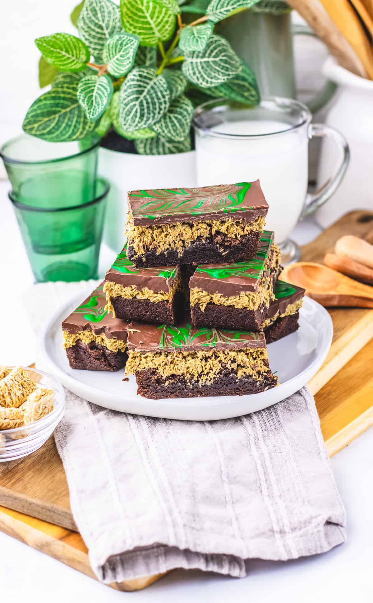 A plate of chocolate layered dessert bars with a green swirl on top, made using a rich Dubai Chocolate Brownie recipe, sits next to a glass of milk and green glasses.