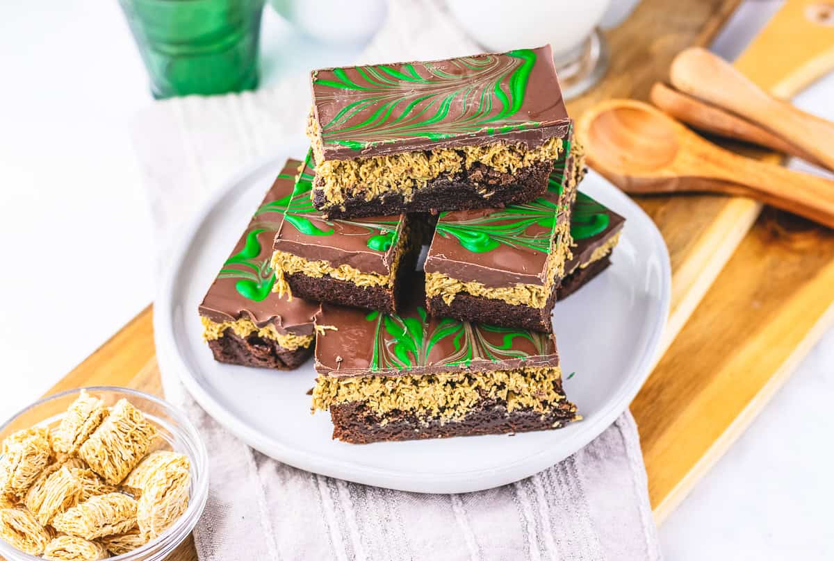 Chocolate and green marbled layered dessert bars stacked on a plate, reminiscent of a classic Dubai Chocolate Brownie, with wooden spoons and cereal nearby.