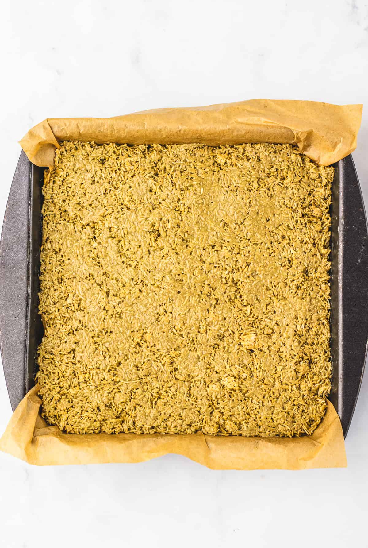 A square baking pan lined with parchment paper, filled with an unbaked oat mixture for a delicious Dubai Chocolate Brownie, sits on a white surface.