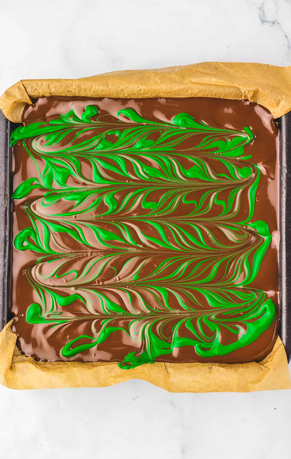 A square tray of Dubai Chocolate Brownie with green swirled patterns on top, lined with parchment paper.