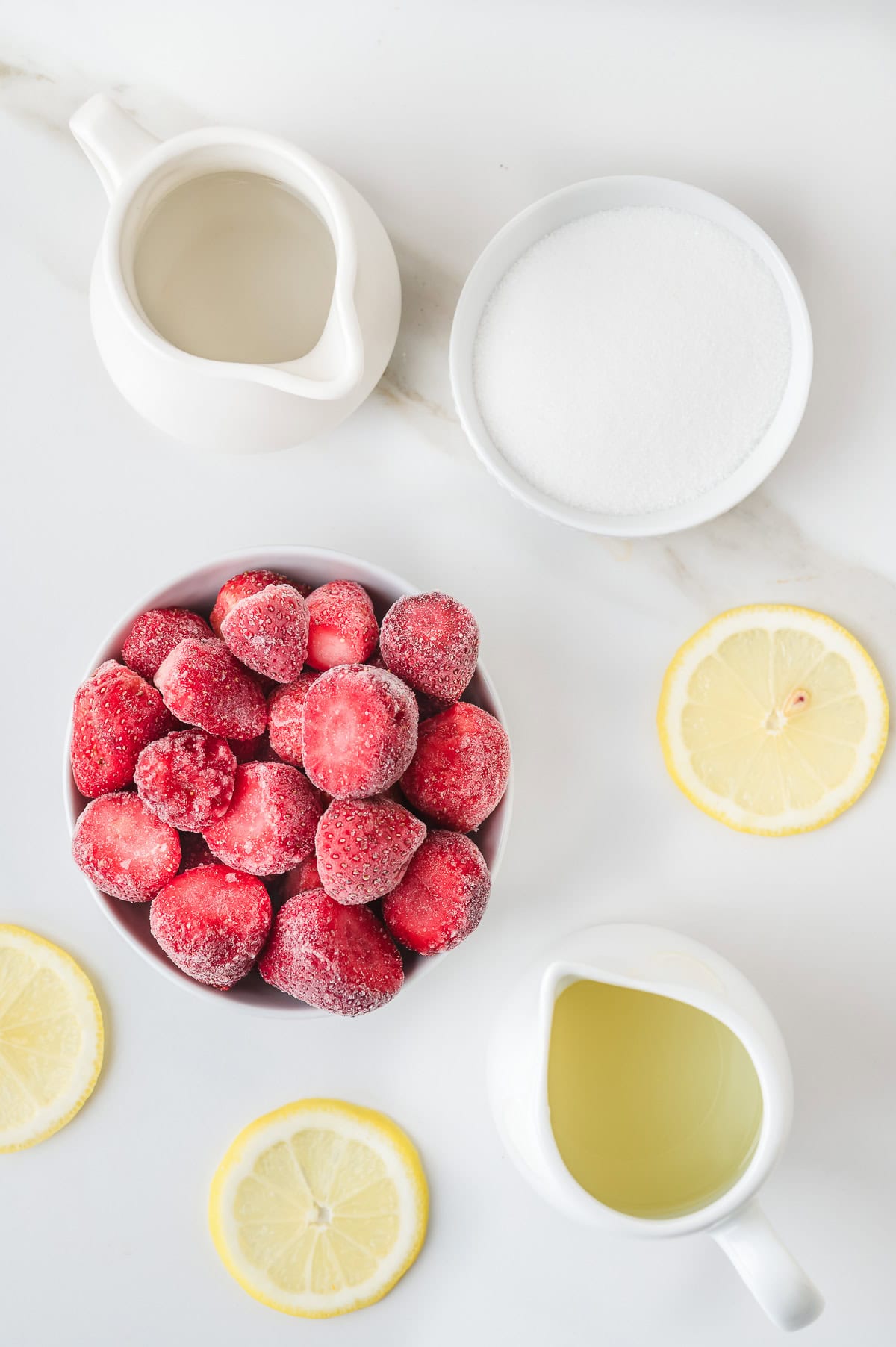 Bowl of frozen strawberries with sugar, lemon slices, water, and syrup in separate containers on a white surface&mdash;perfect for making a refreshing Frozen Strawberry Lemonade.