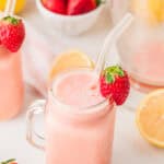 A pink smoothie in a mason jar with a glass straw, garnished with a strawberry, surrounded by lemons and strawberries—a refreshing nod to classic Frozen Strawberry Lemonade.
