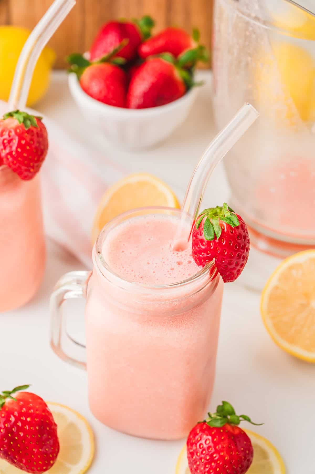 A pink smoothie in a mason jar with a glass straw, garnished with a strawberry, surrounded by lemons and strawberries&mdash;a refreshing nod to classic Frozen Strawberry Lemonade.