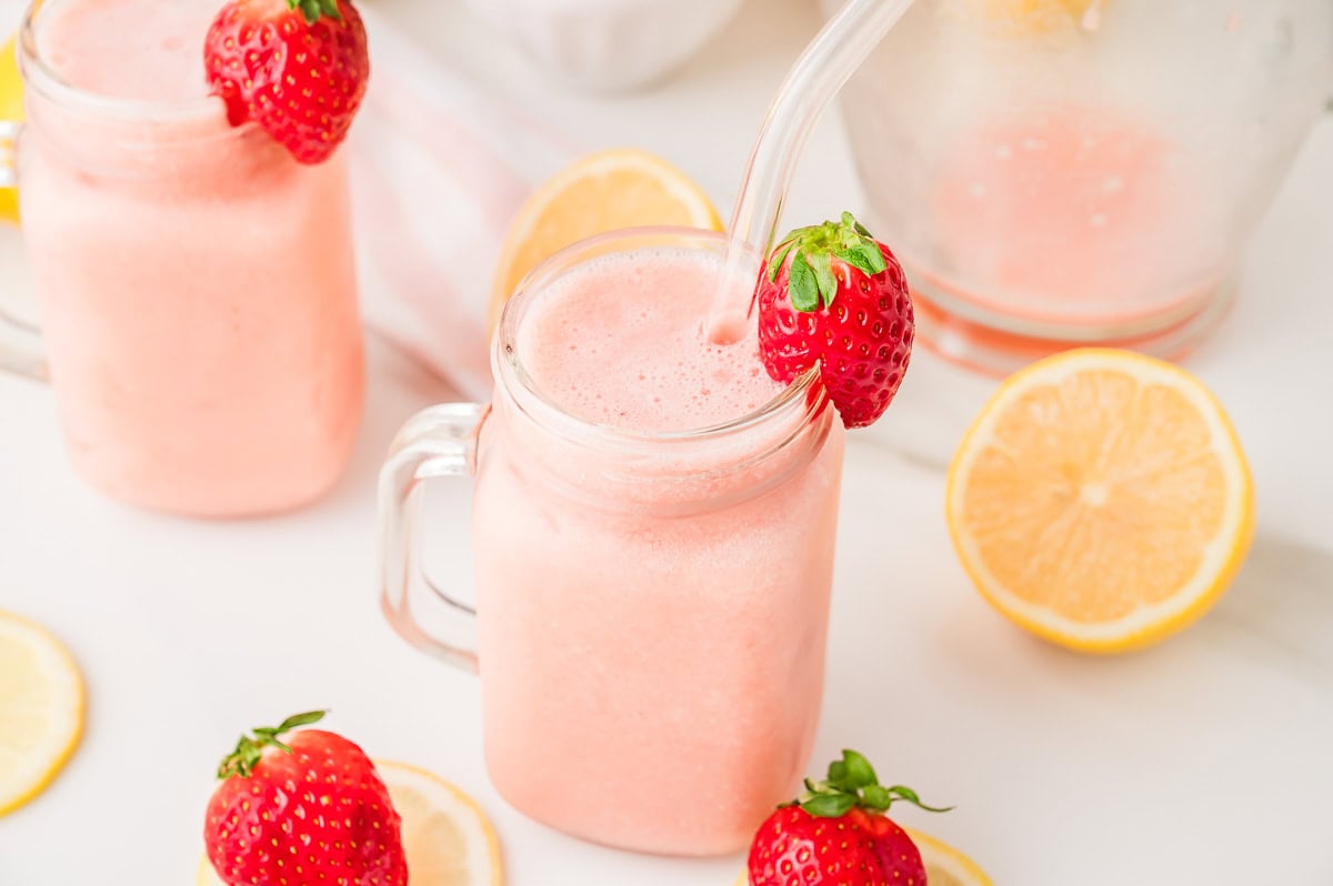 Glass jars of pink frozen strawberry lemonade with whole strawberries and lemon slices on a white surface.