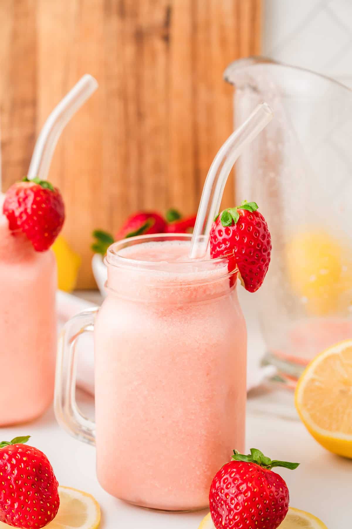 A pink frozen strawberry lemonade smoothie in a mason jar with a glass straw, garnished with a strawberry and surrounded by lemons.