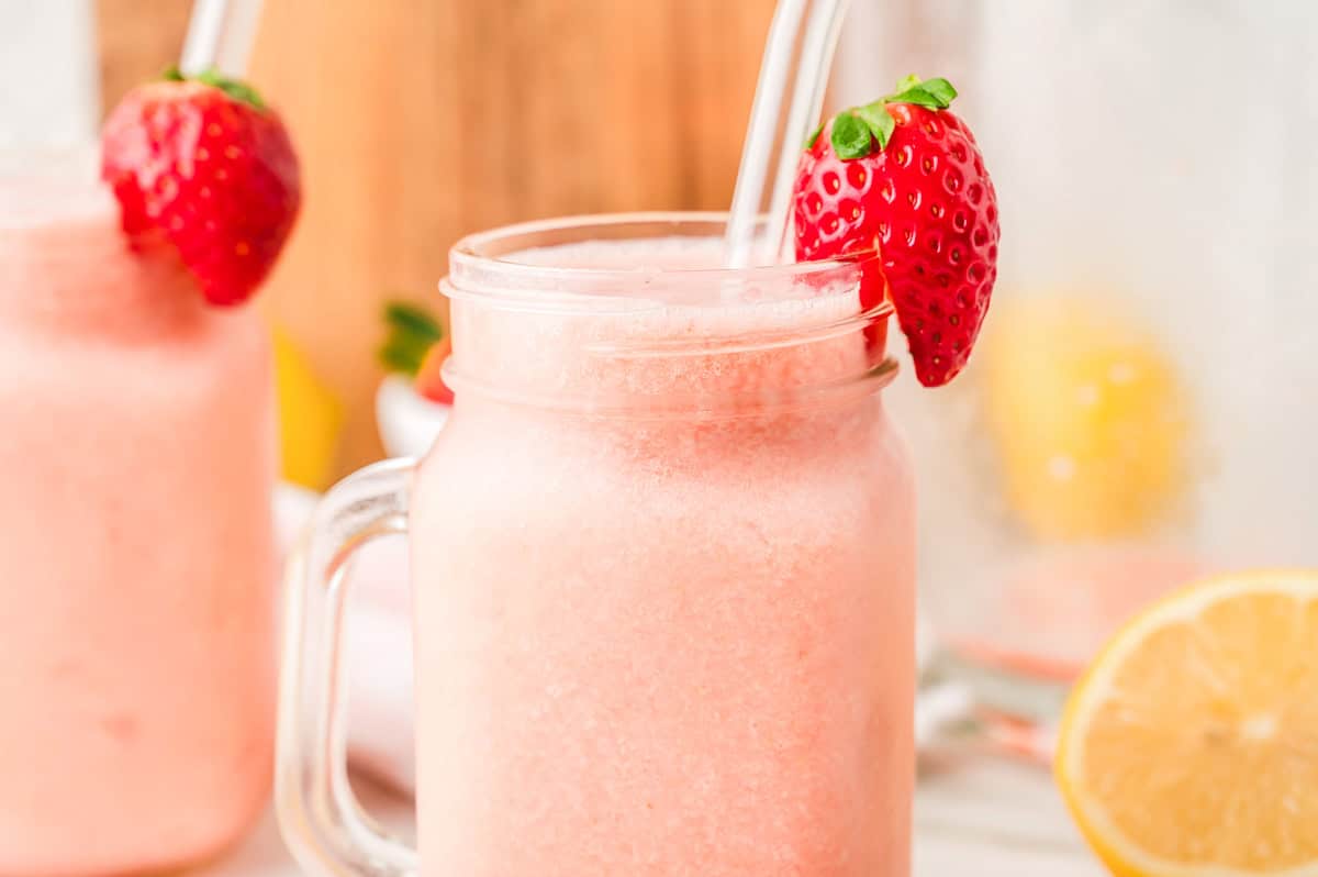 A pink strawberry smoothie in a mason jar with a clear straw and a fresh strawberry on the rim, perfect for those who love a refreshing frozen strawberry lemonade.