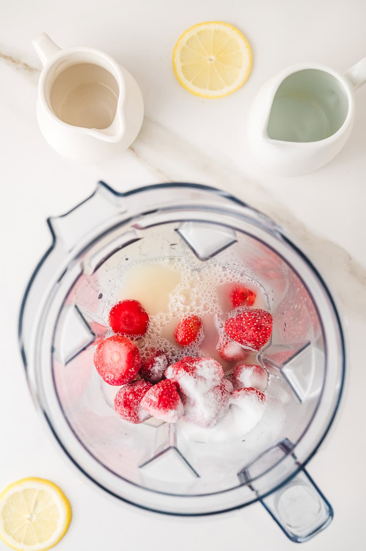 Top view of a blender with strawberries, sugar, and liquid, surrounded by lemon slices and pitchers&mdash;perfect for making a refreshing Frozen Strawberry Lemonade.