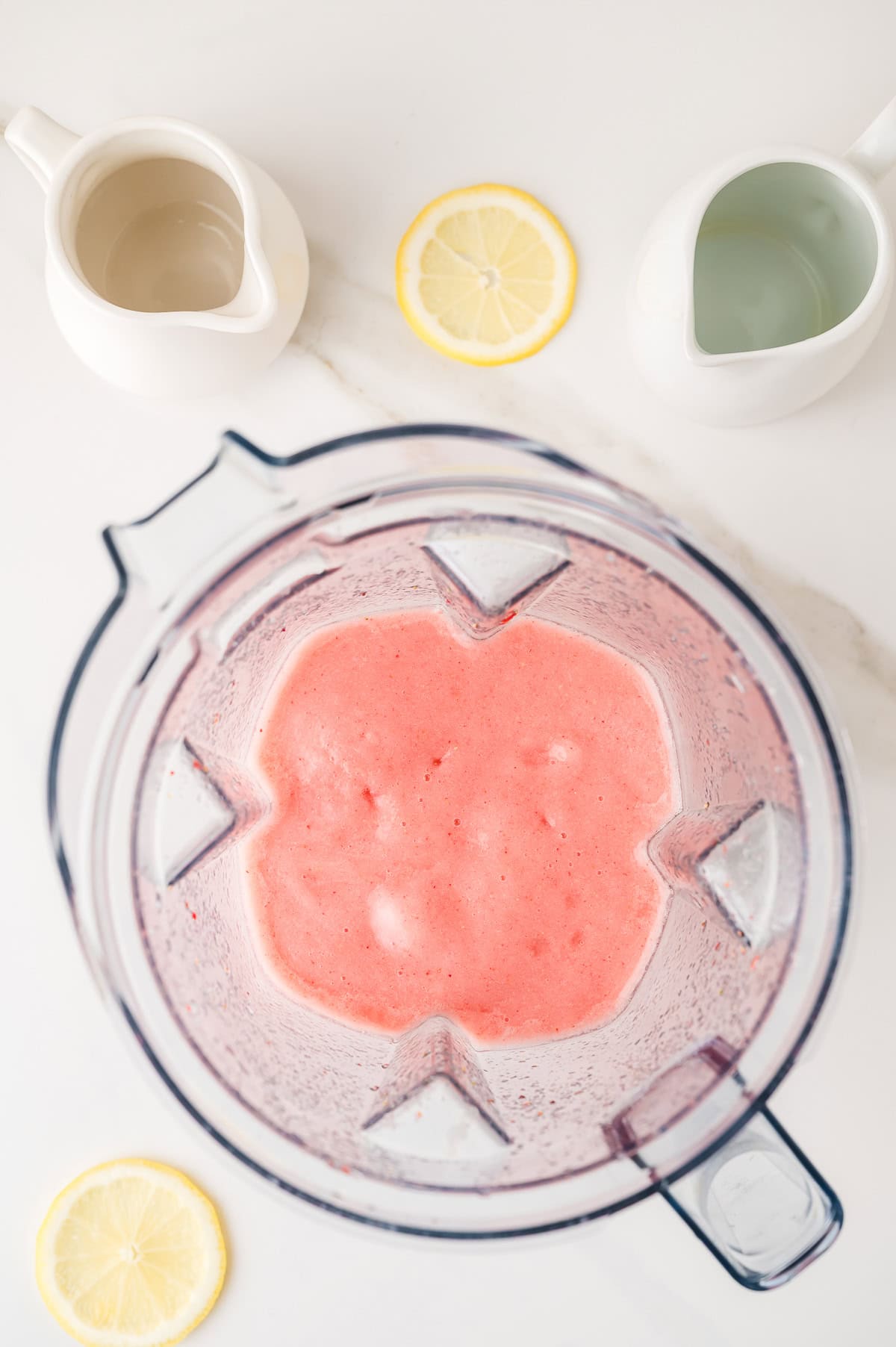 Top view of a blender with pink smoothie, two lemon slices, and two white pitchers on a white surface&mdash;perfect inspiration for a Frozen Strawberry Lemonade.