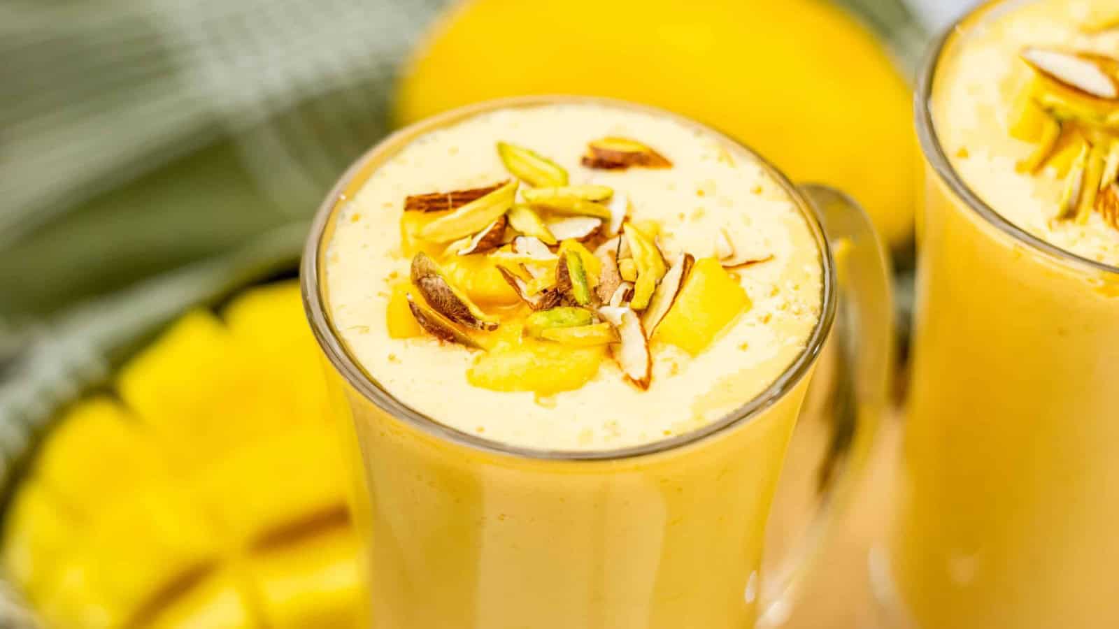 A glass of mango lassi topped with chopped nuts and mango pieces, with fresh mango in the background.