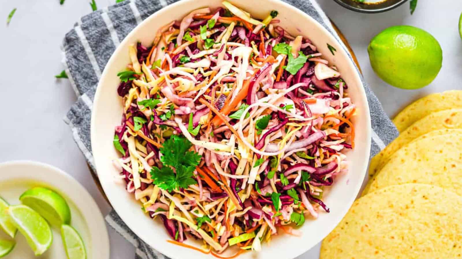 Bowl of colorful coleslaw with shredded cabbage, carrots, and cilantro, next to lime wedges and tortillas.