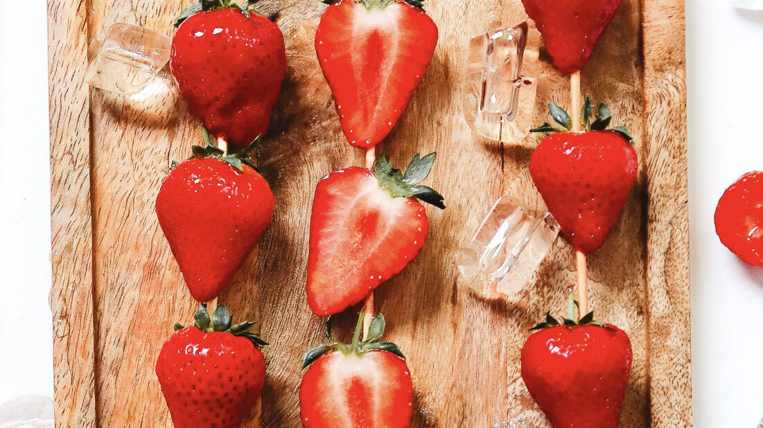 Three skewers with whole and halved strawberries and ice cubes on a wooden board, viewed from above&mdash;perfect inspiration for your next Strawberry Tanghulu or Tanghulu recipe.