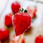 A shiny, glazed strawberry on a wooden stick showcases the classic Strawberry Tanghulu, with blurred strawberries in the background.