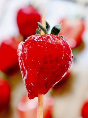 A shiny, glazed strawberry on a wooden stick showcases the classic Strawberry Tanghulu, with blurred strawberries in the background.