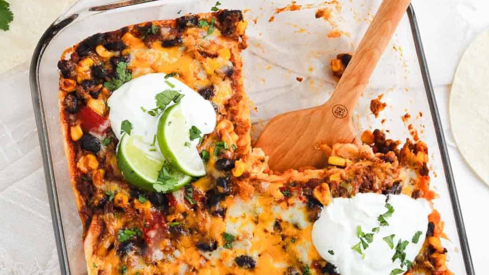 A casserole topped with melted cheese, black beans, corn, sour cream, lime slices, and cilantro in a glass dish.