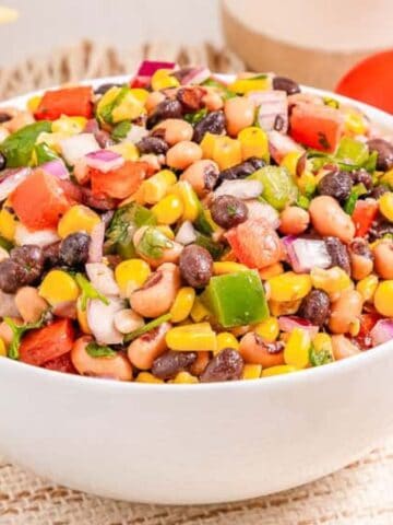 A bowl of colorful bean and corn salad with diced vegetables, next to lime halves, a tomato, and tortilla chips.