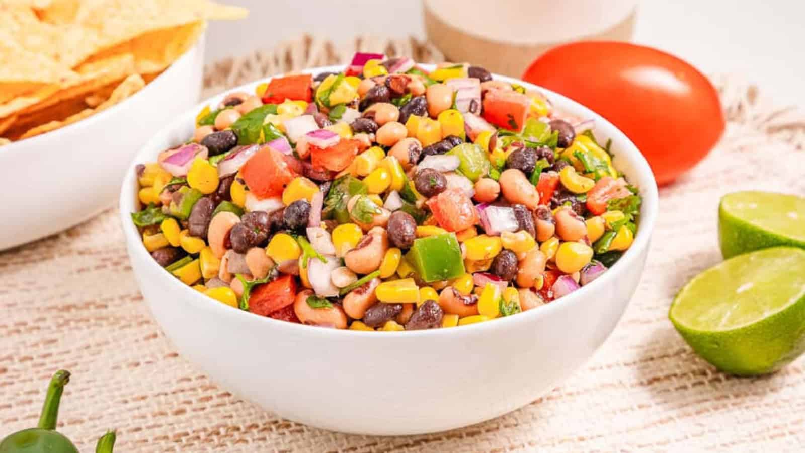 A bowl of colorful bean and corn salsa with chopped tomato, onion, cilantro, and lime on the side.
