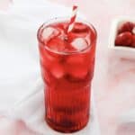 Tall glass of red iced drink with a striped straw, on a white cloth with strawberries in the background&mdash;a perfect Starbucks Copycat Strawberry Acai Refresher.