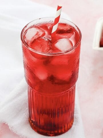 Tall glass of red iced drink with a striped straw, on a white cloth with strawberries in the background—a perfect Starbucks Copycat Strawberry Acai Refresher.