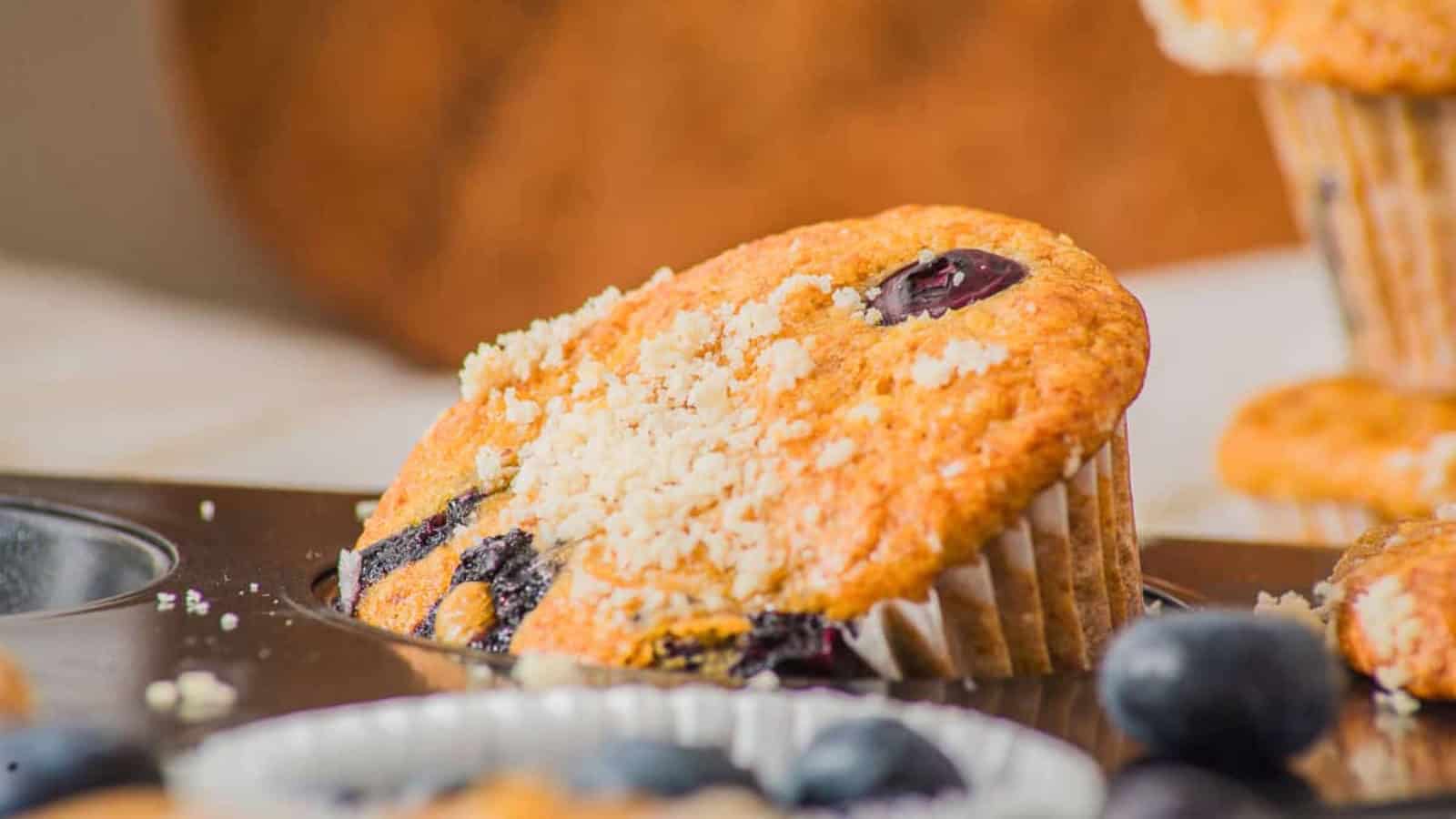 Blueberry muffin with crumb topping in a muffin tin, surrounded by fresh blueberries.