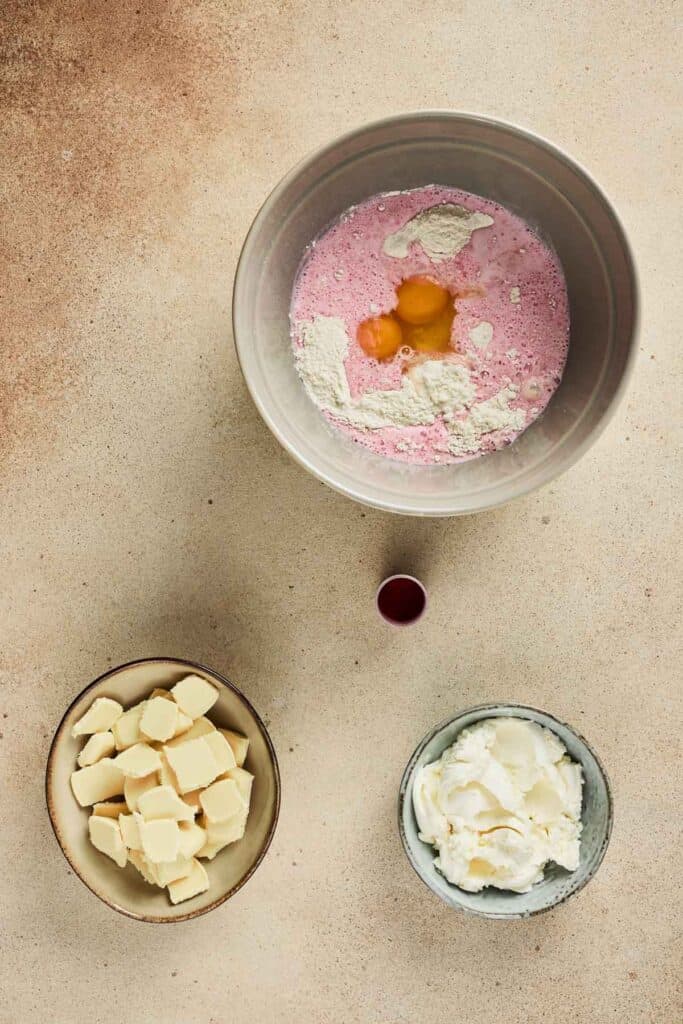 A bowl with eggs, flour, and pink liquid&mdash;perfect for a Strawberry Cake Balls Recipe&mdash;sits beside bowls of butter, cream, and a small cup of red liquid.
