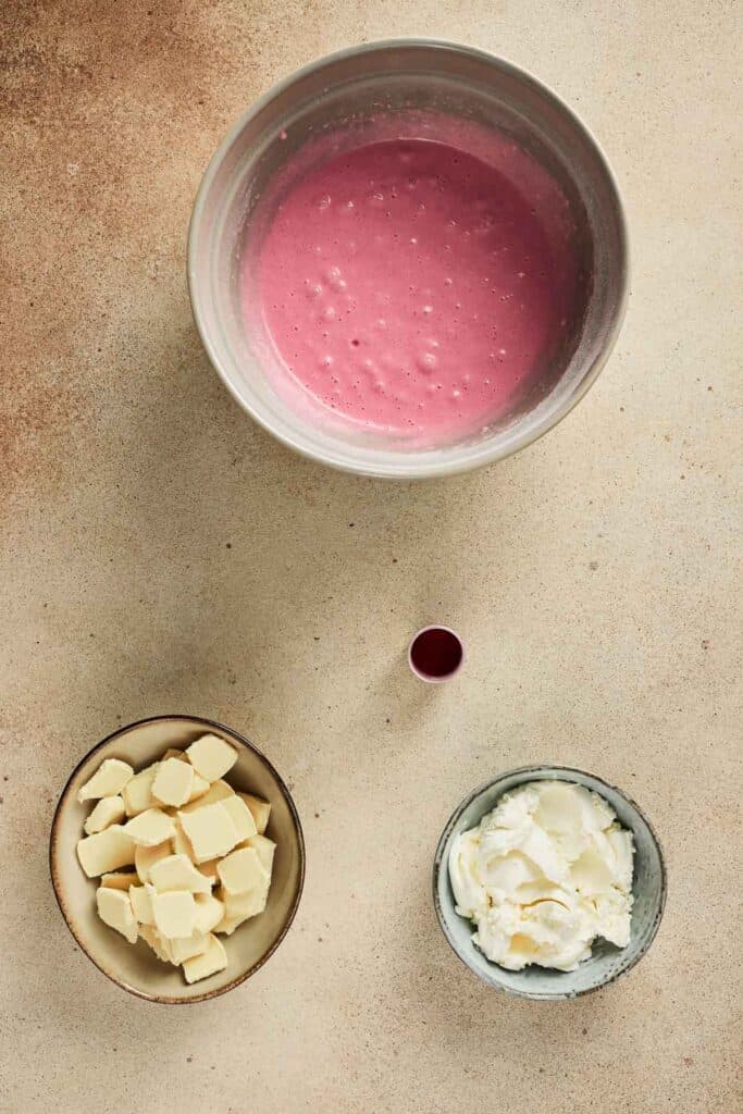 Three bowls with pink batter for Strawberry Cake, cubed butter, and cream cheese, plus a small cup of red liquid sit on a beige surface.