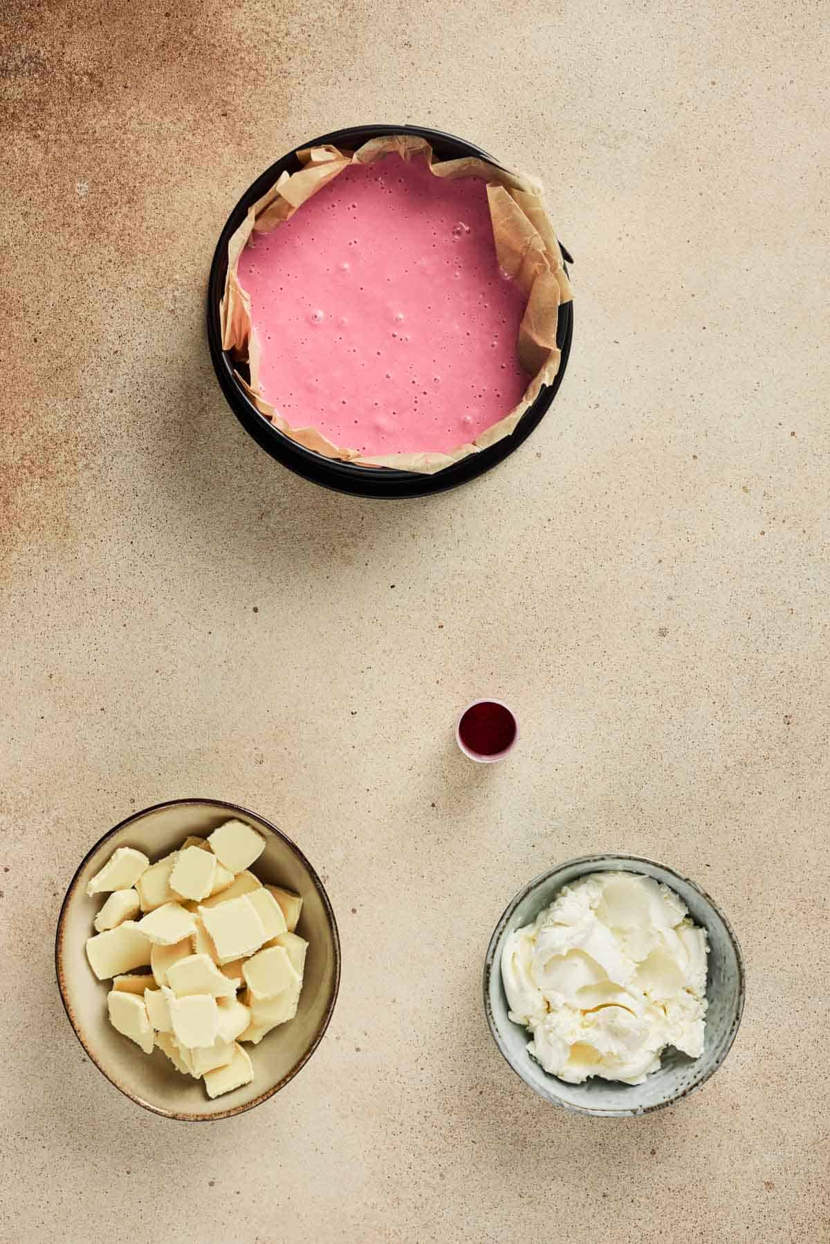 Three bowls with pink mixture for a Strawberry Cake Balls recipe, chopped white chocolate, whipped cream, and a small cup of red liquid are arranged on a tan surface.