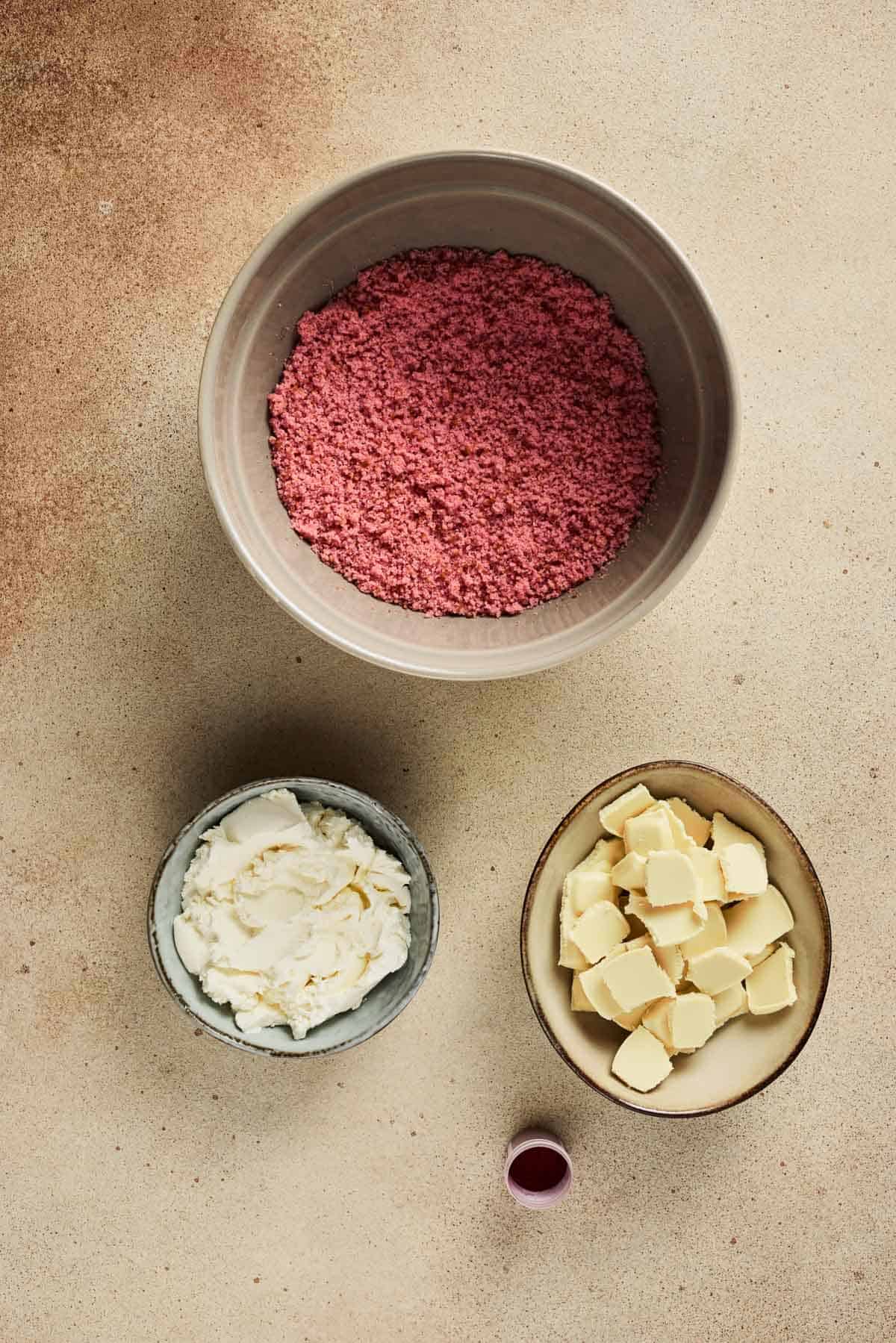 Three bowls with pink crumbs, white cream, and butter cubes, plus a small cup of pink liquid on a beige surface&mdash;perfect ingredients for a delicious Strawberry Cake Balls recipe.