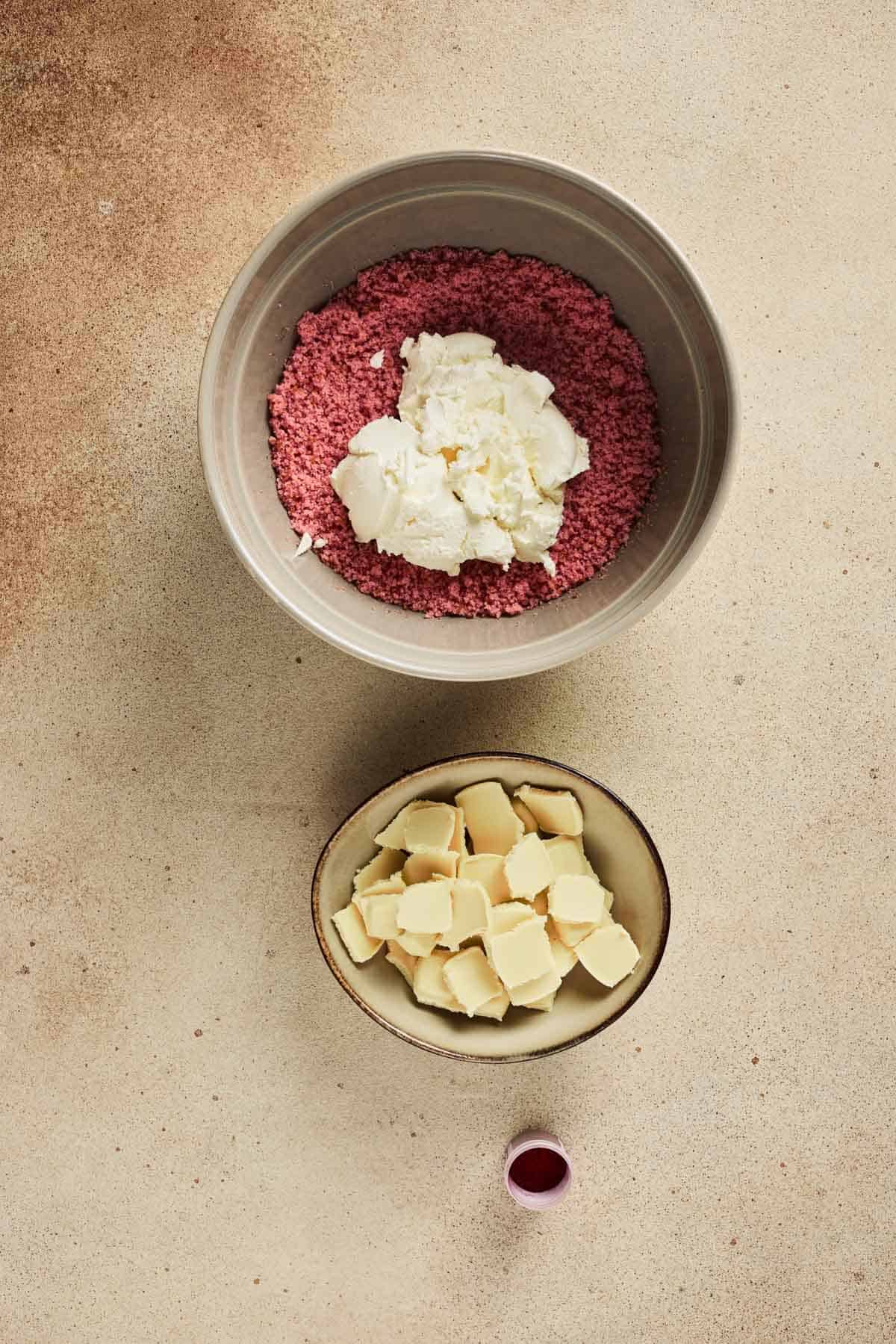 Two bowls with pink crumbs for Strawberry Cake Balls, cream cheese, white chocolate chunks, and a small cup of red liquid sit on a tan surface.