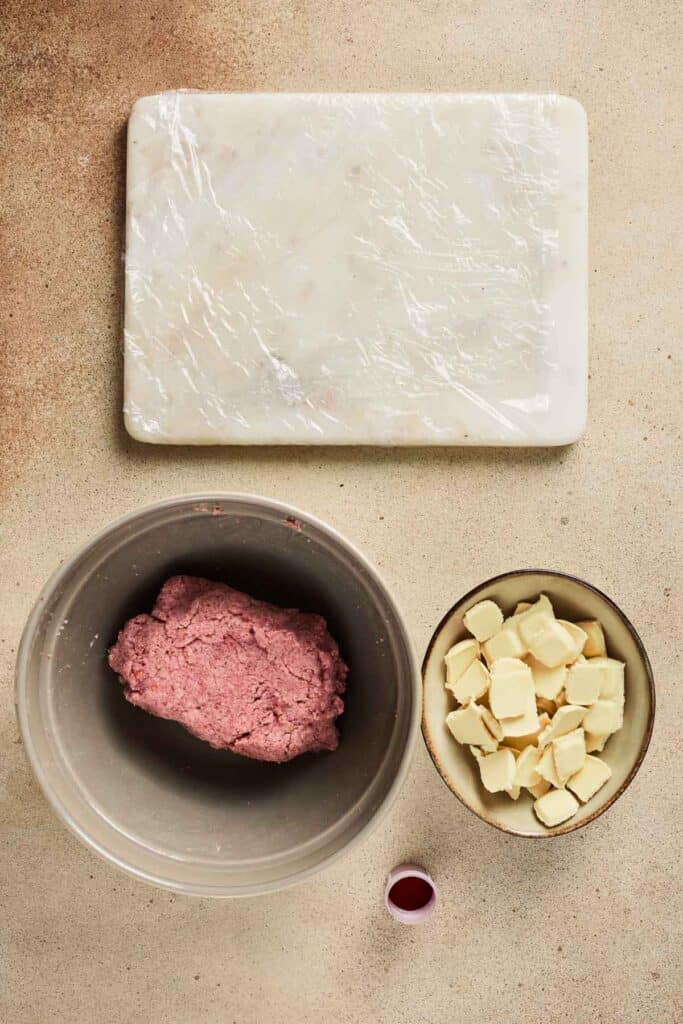 A bowl of pink dough for Strawberry Cake Balls, a bowl of butter pieces, a small cup of red liquid, and a wrapped board rest on the counter&mdash;essential ingredients for this delicious Strawberry Dessert.