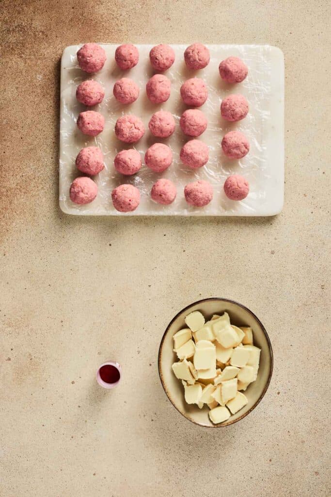 Rows of pink Strawberry Cake Balls on a tray, a bowl of white chocolate chunks, and a small cup of red liquid ready for the Strawberry Cake Balls Recipe.