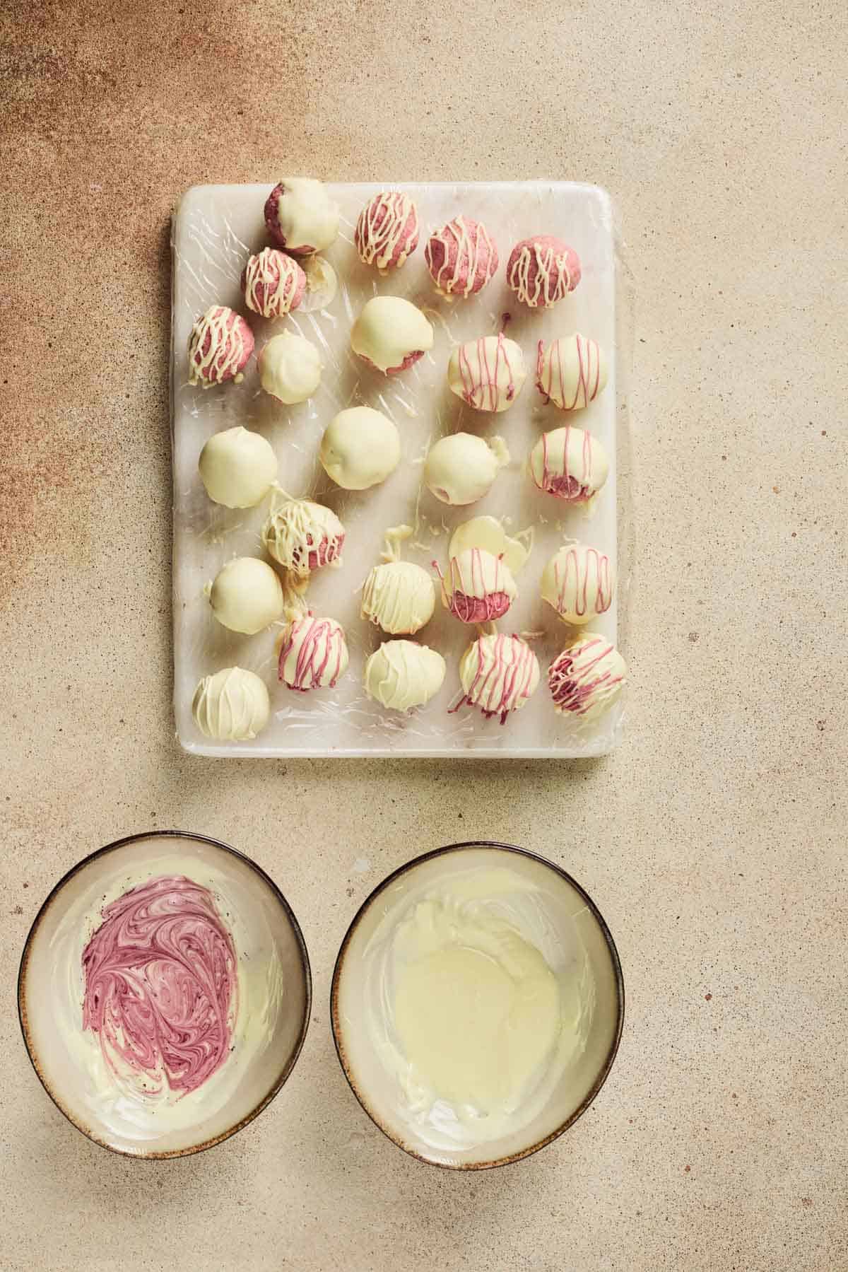 Twenty decorated strawberry cake balls on a tray, with two bowls of melted chocolate&mdash;one pink and one white&mdash;below, make this a delightful strawberry dessert display.
