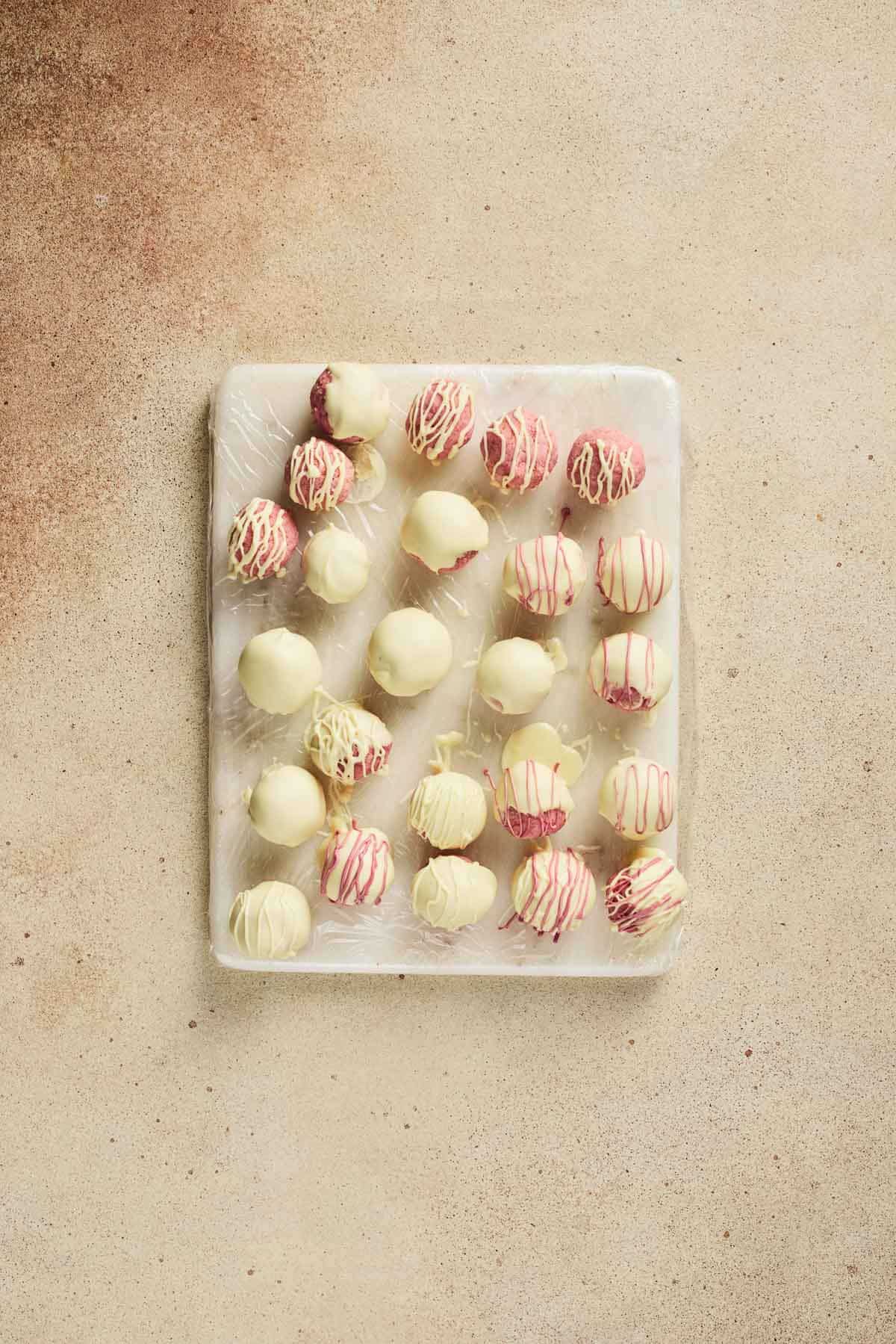 A tray of white chocolate-covered Strawberry Cake Balls, some drizzled with red and white chocolate, sits on a beige surface&mdash;an irresistible strawberry dessert for any occasion.