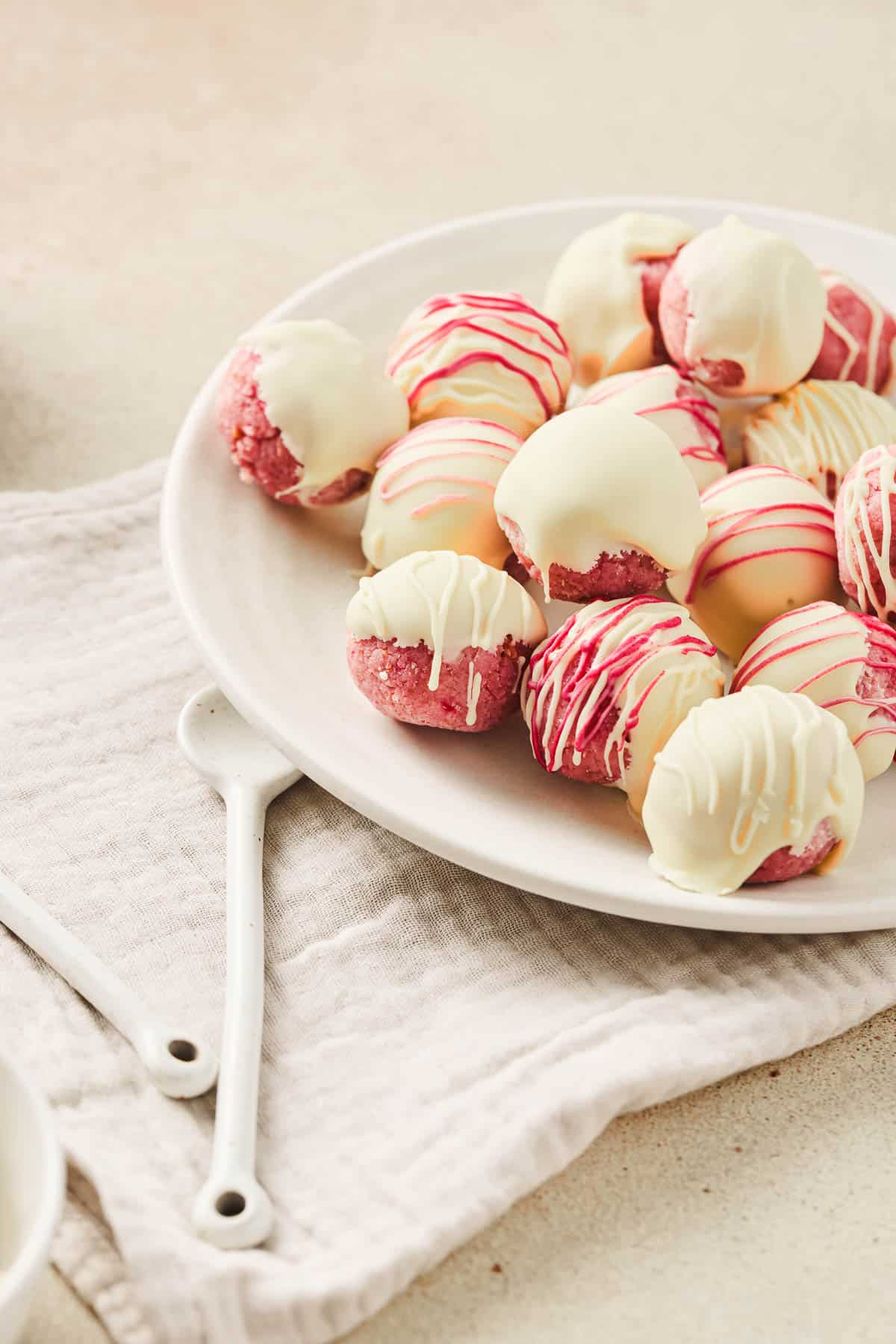 A plate of pink cookies, inspired by a Strawberry Cake Balls recipe, covered in white chocolate and pink drizzle, rests on a white napkin with two white spoons.