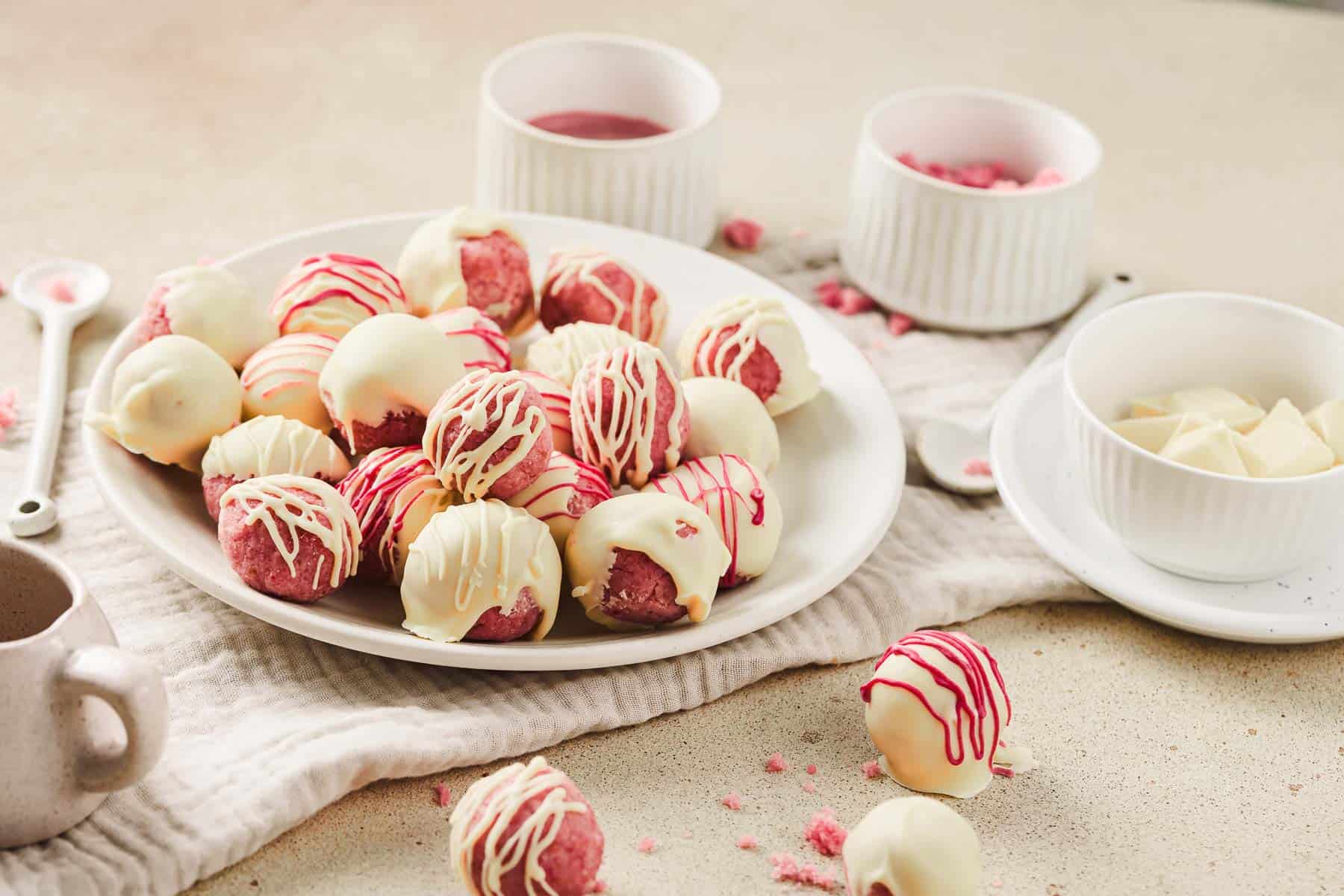 A plate of Strawberry Cake Balls drizzled with white and pink chocolate, surrounded by bowls of chocolate and toppings&mdash;an irresistible strawberry dessert inspired by your favorite cake balls recipe.