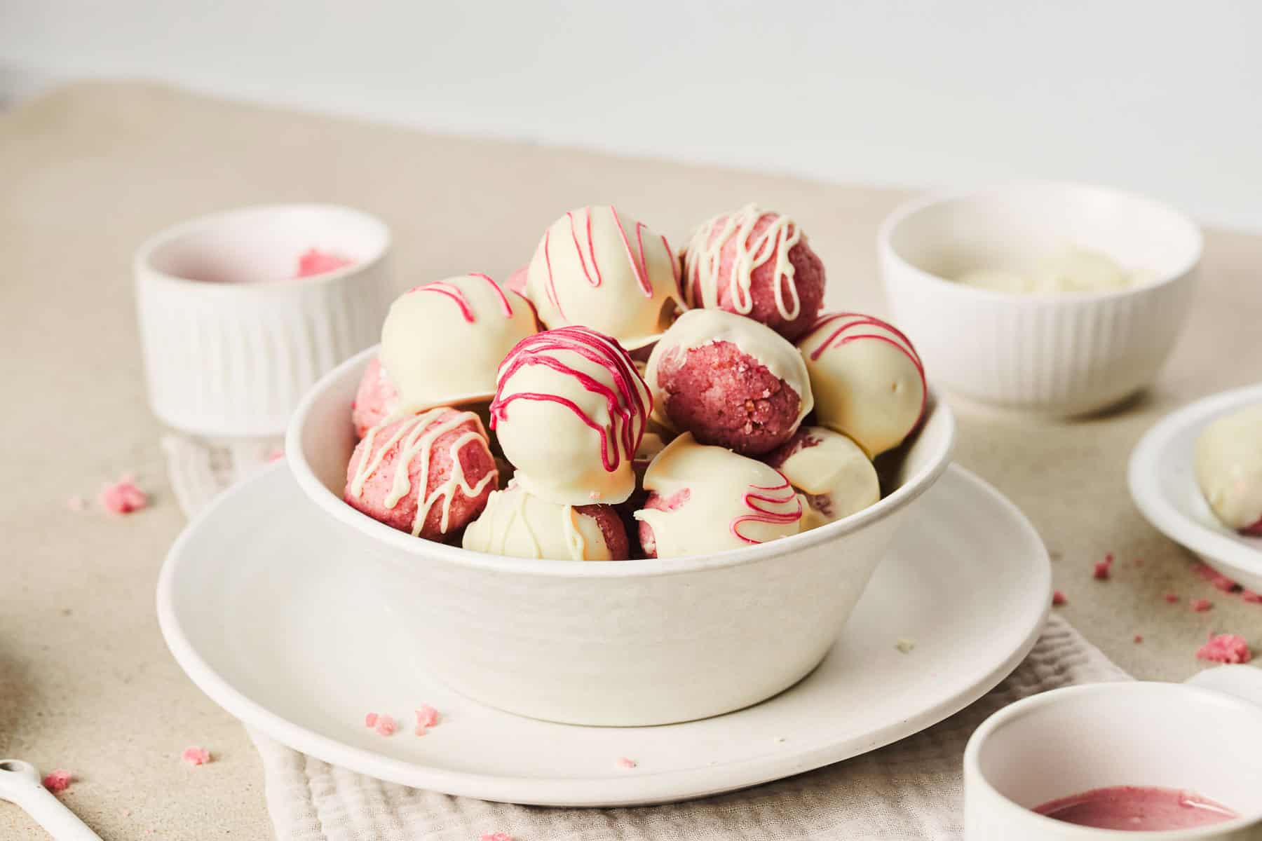 A bowl of pink and white Strawberry Cake Balls drizzled with pink icing, set on a white plate with bowls nearby&mdash;an irresistible strawberry dessert.