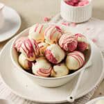 A bowl of pink Strawberry Cake Balls coated in white chocolate, some drizzled with red, on a white plate with a spoon—a delightful strawberry dessert.