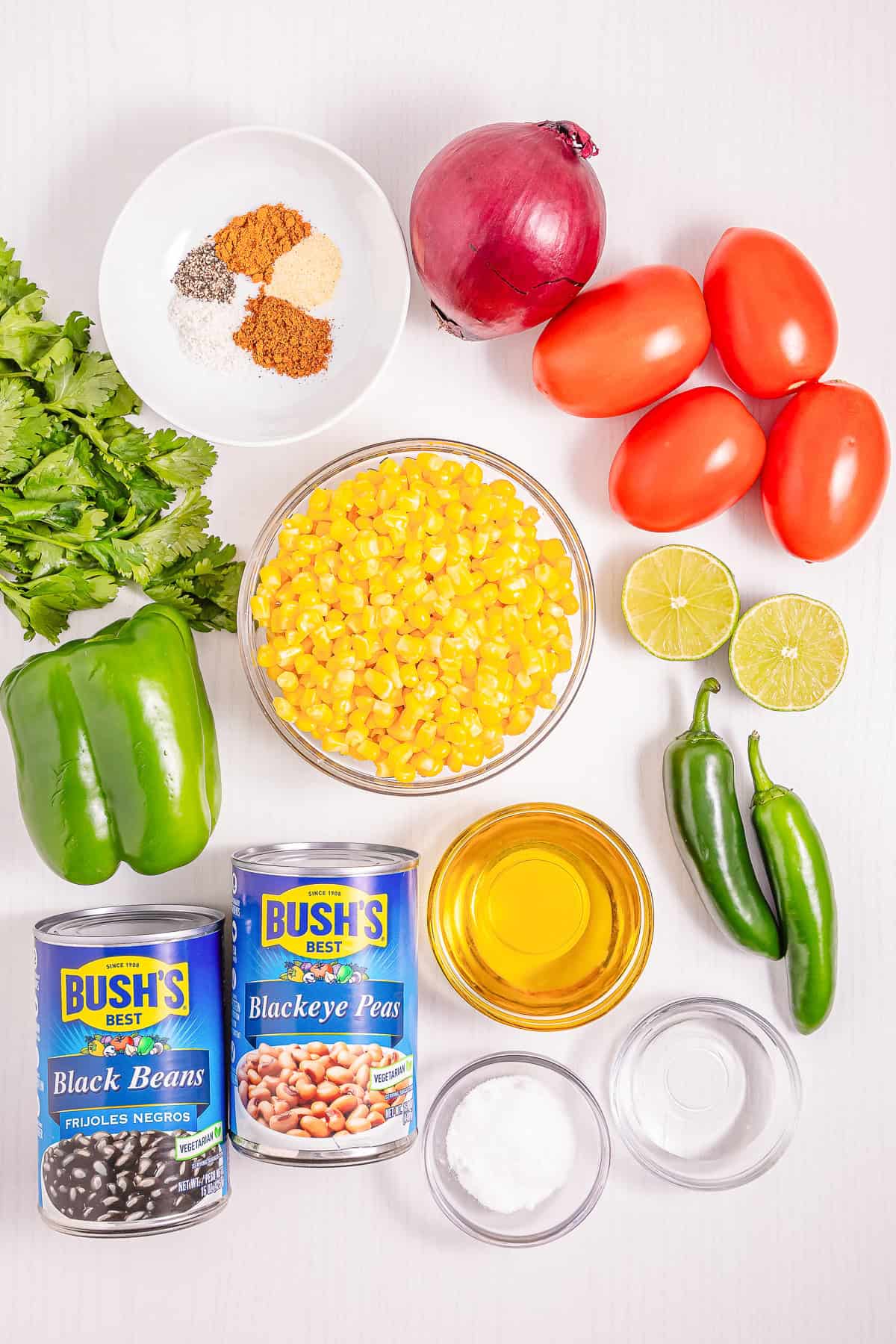 Top-down view of fresh vegetables, canned beans, lime, spices, and oil on a white surface&mdash;perfect ingredients for a vibrant Cowboy Caviar Recipe.
