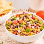 A bowl of colorful cowboy caviar salad with corn, tomatoes, and herbs, served next to tortilla chips and fresh vegetables.